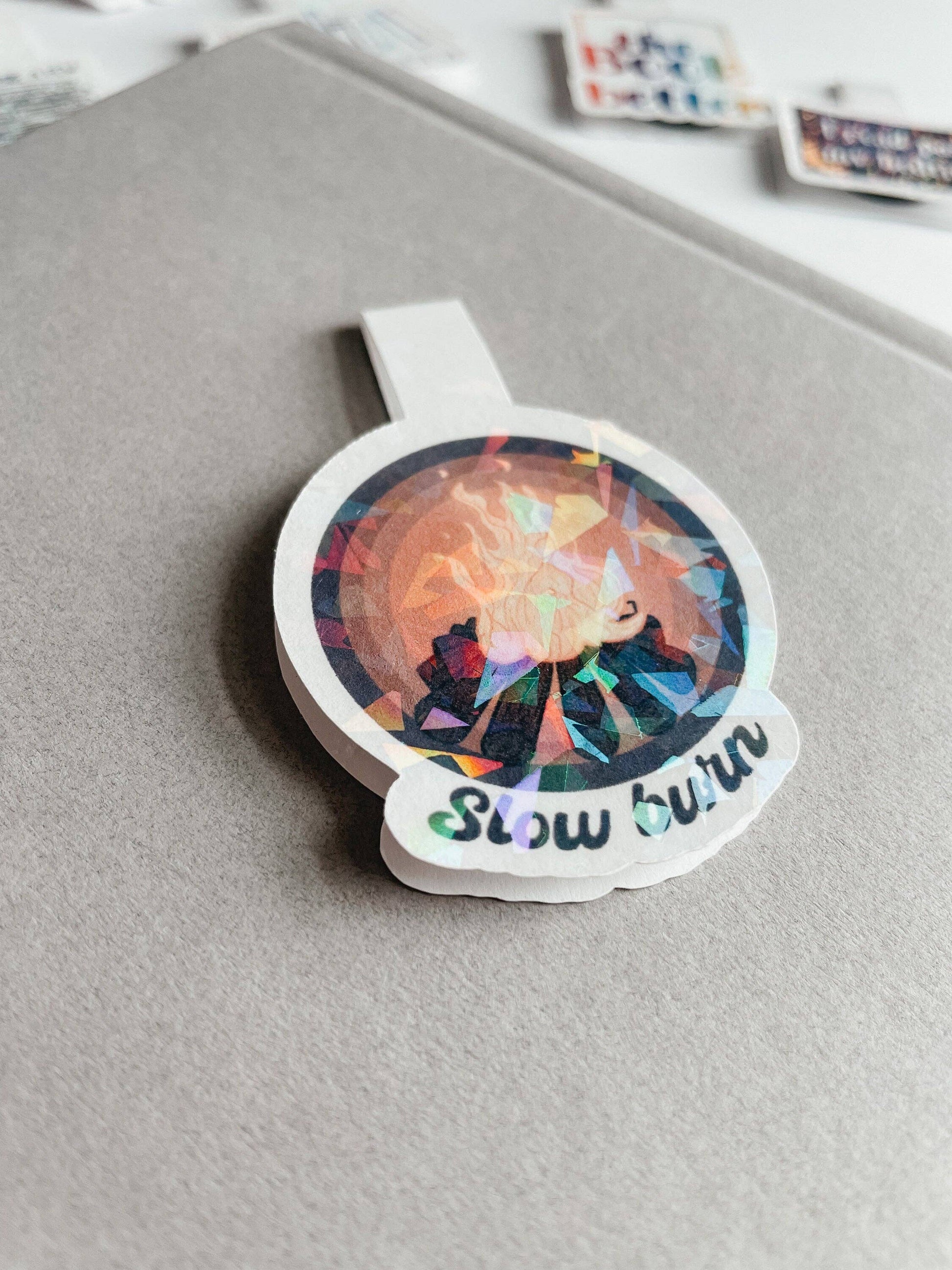 Holographic sticker with a colorful design and 'Slow burn' text on a gray surface