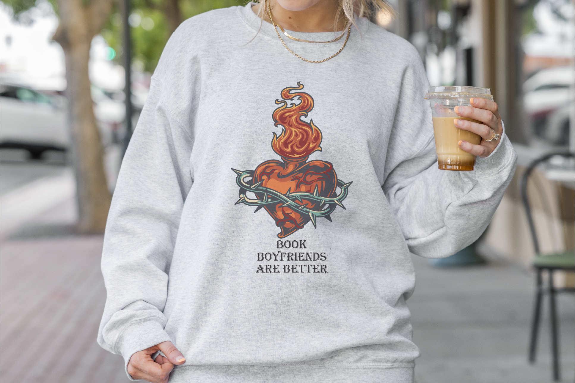 Person wearing a gray sweatshirt with a graphic design and text, holding a coffee cup outdoors.